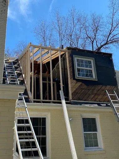 An exterior view of the same second-story home addition during the construction phase, showing exposed wooden wall framing, roof structure, and a ladder leaning against the house.