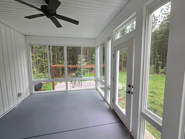 An interior view of a sunroom, featuring grey flooring, white walls, and a black ceiling fan, with large windows overlooking a backyard with trees and a trellis.