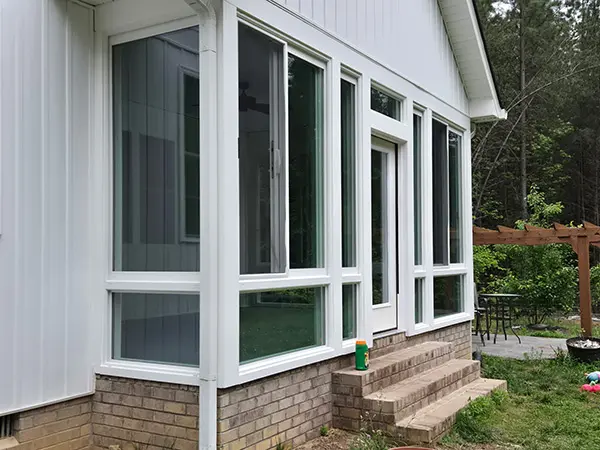 An exterior view of a modern white sunroom built onto a house with white vertical siding and a brick foundation. Multiple large windows and a glass-paneled door are visible.