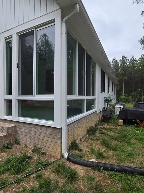 An exterior, side-corner view of a sunroom, showing numerous windows and a black gutter drainage pipe running along the ground next to the brick foundation.