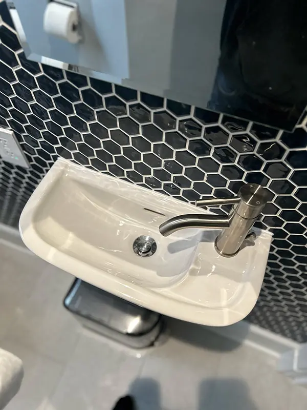 A top-down view of a small, modern wall-mounted bathroom sink with a brushed nickel faucet, set against a dark hexagonal tile wall.