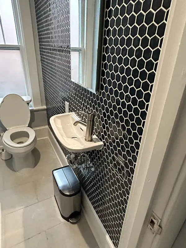 An angled interior shot of a small modern bathroom featuring the dark hexagonal tile wall, the wall-mounted sink, a silver trash can, and a toilet.