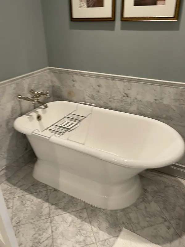 A white freestanding soaking tub positioned next to a half-wall covered in light gray marble tile.