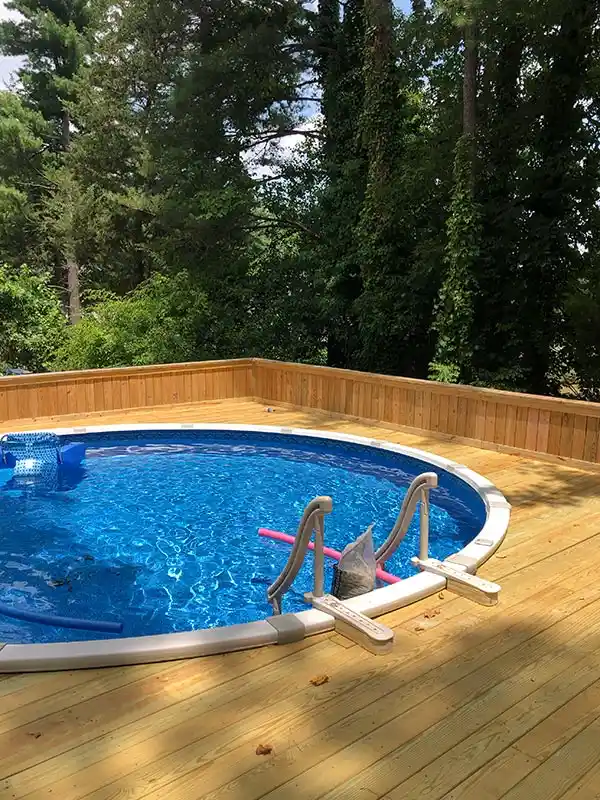 A spacious custom-built wooden deck surrounding a large, round above-ground swimming pool, nestled in a lush, wooded backyard.