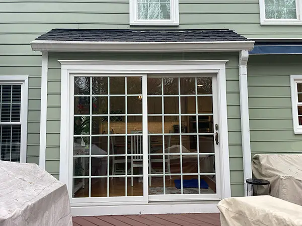 Exterior view of a white sliding glass door with a grid pattern, installed against light green horizontal siding under a small shingled awning, leading out onto a wooden deck with covered patio furniture.