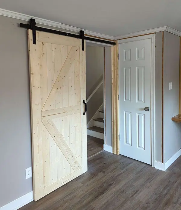 A light wood sliding barn door with black metal track hardware, partially open to reveal a hallway with a staircase and a standard white interior door.