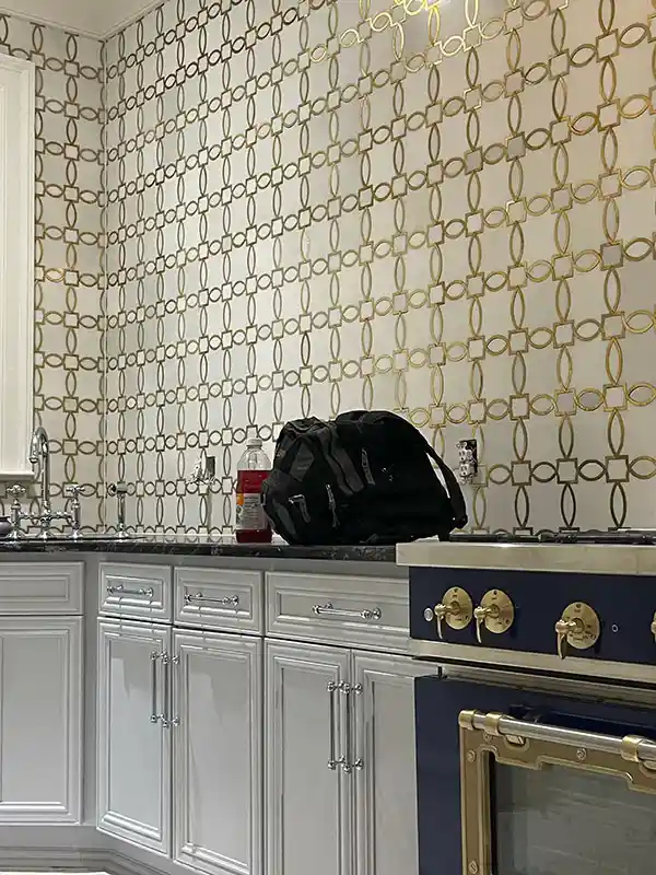 An interior kitchen shot featuring white cabinetry with silver hardware, a dark countertop, and a striking white backsplash with a gold interlocking geometric pattern, situated next to a navy blue stove with gold accents.