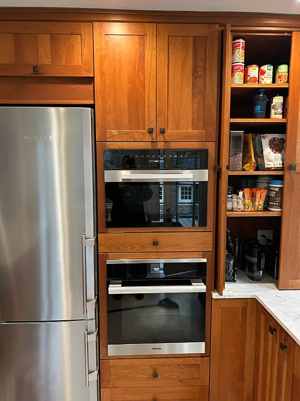 An eye-level close-up shot of built-in stainless steel appliances, including an oven and a microwave, surrounded by medium-wood cabinets and an open pantry cupboard filled with items.