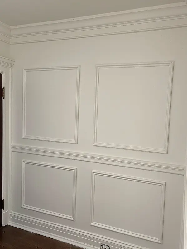 An interior white wall featuring elegant architectural details, including traditional wainscoting and large, raised picture frame molding panels.
