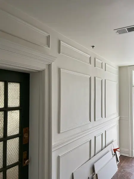 An interior shot of a wall featuring ornate white wainscoting and large panel moldings, with a dark-framed, multi-pane glass door to the left.