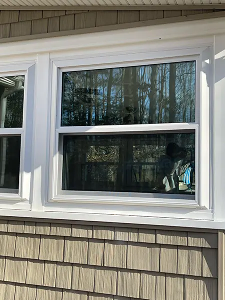 A close-up exterior view of a modern white-framed window set against tan shingle siding, with the glass reflecting a dense forest of bare trees.