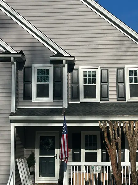 The front exterior of a house with light gray horizontal siding, featuring two upper-story windows with black shutters and a white-railed porch where an American flag is displayed near the front door.